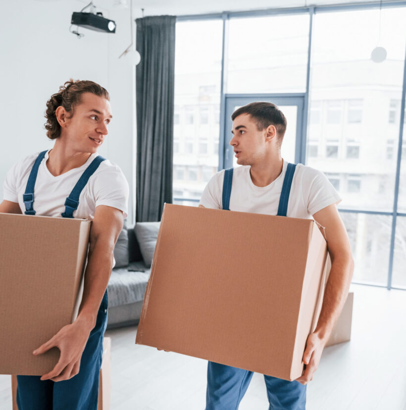 Busy day. Two young movers in blue uniform working indoors in the room.